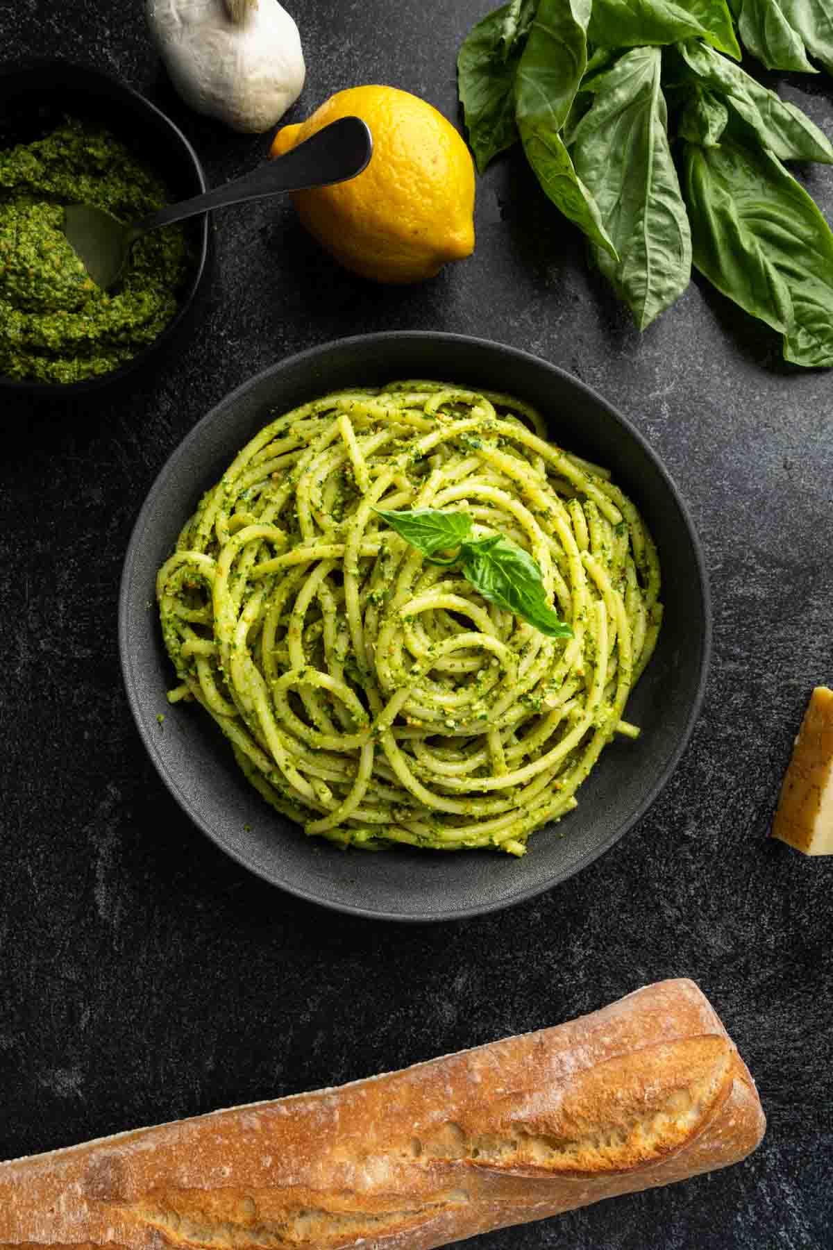 A big bowl of spaghetti al pesto surrounded by basil, lemon, pesto and a baguette.