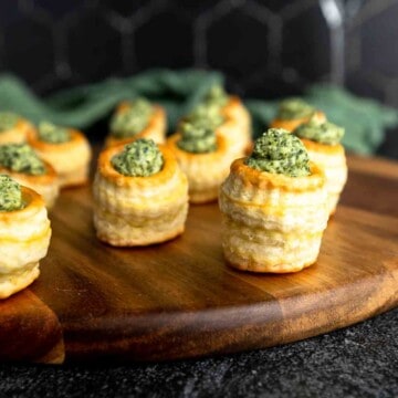 A wooden tray topped with mini vol-au-vent puff pastry cups filled with spinach and artichoke dip.