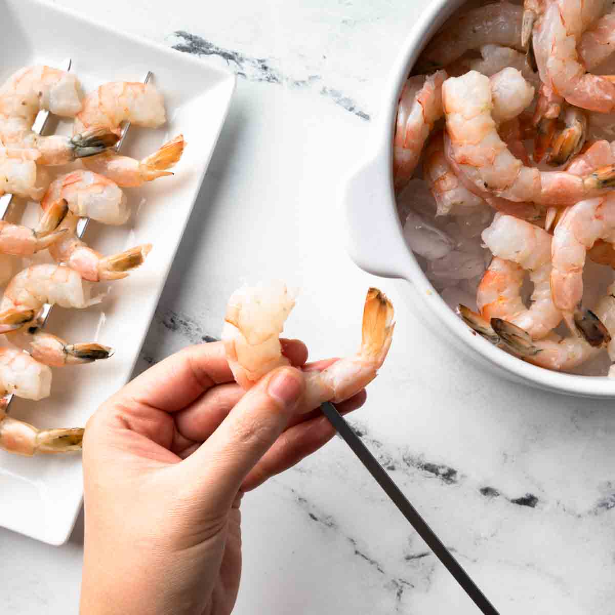 A flat metal skewer being inserted just above the tail of the shrimp. 