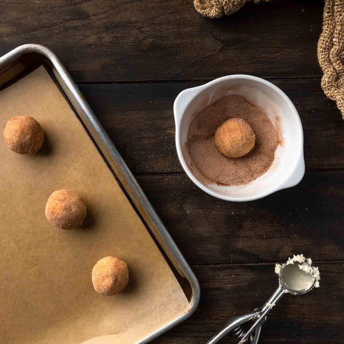 Balls of cookie dough on a sheet tray next to a cookie dough ball in cinnamon sugar. 