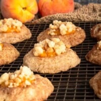 A baking rack full of peach cobbler cookies next to a couple fresh peaches.