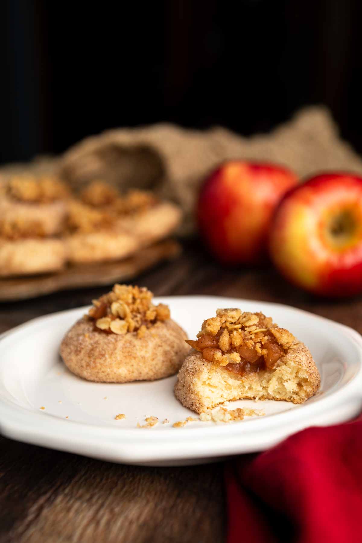 A bitten apple crumble cookie on a white plate with a platter in the background. 