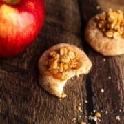 A bitten apple crumble cookie next to a sprinkle of crumbs and a fresh apple.