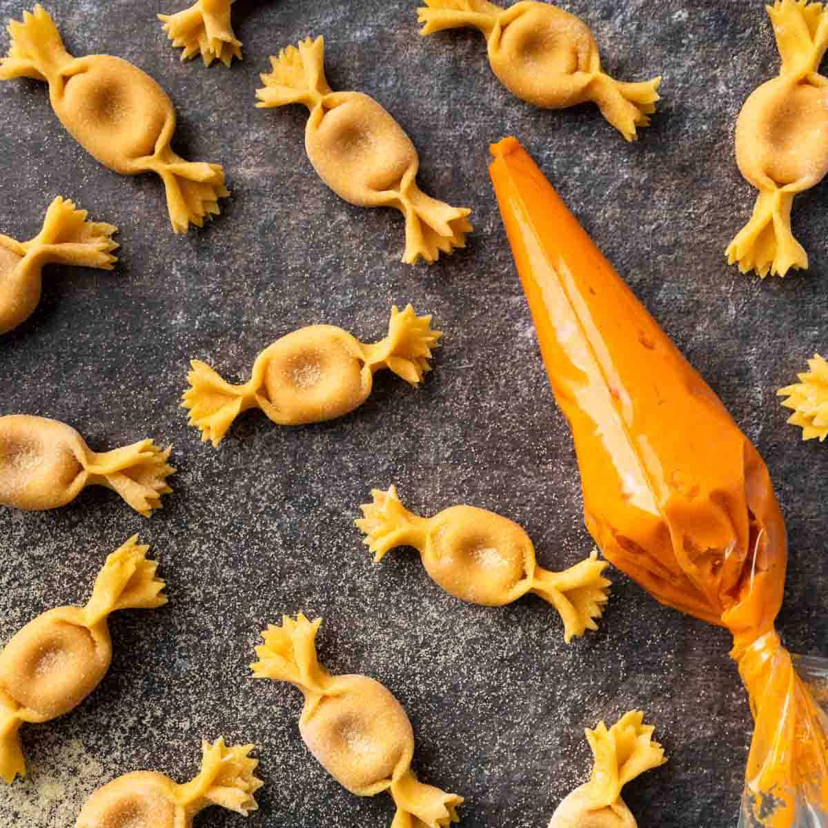 A countertop scattered with handmade caramelle pasta next to a piping bag filled with sweet potato farce. 