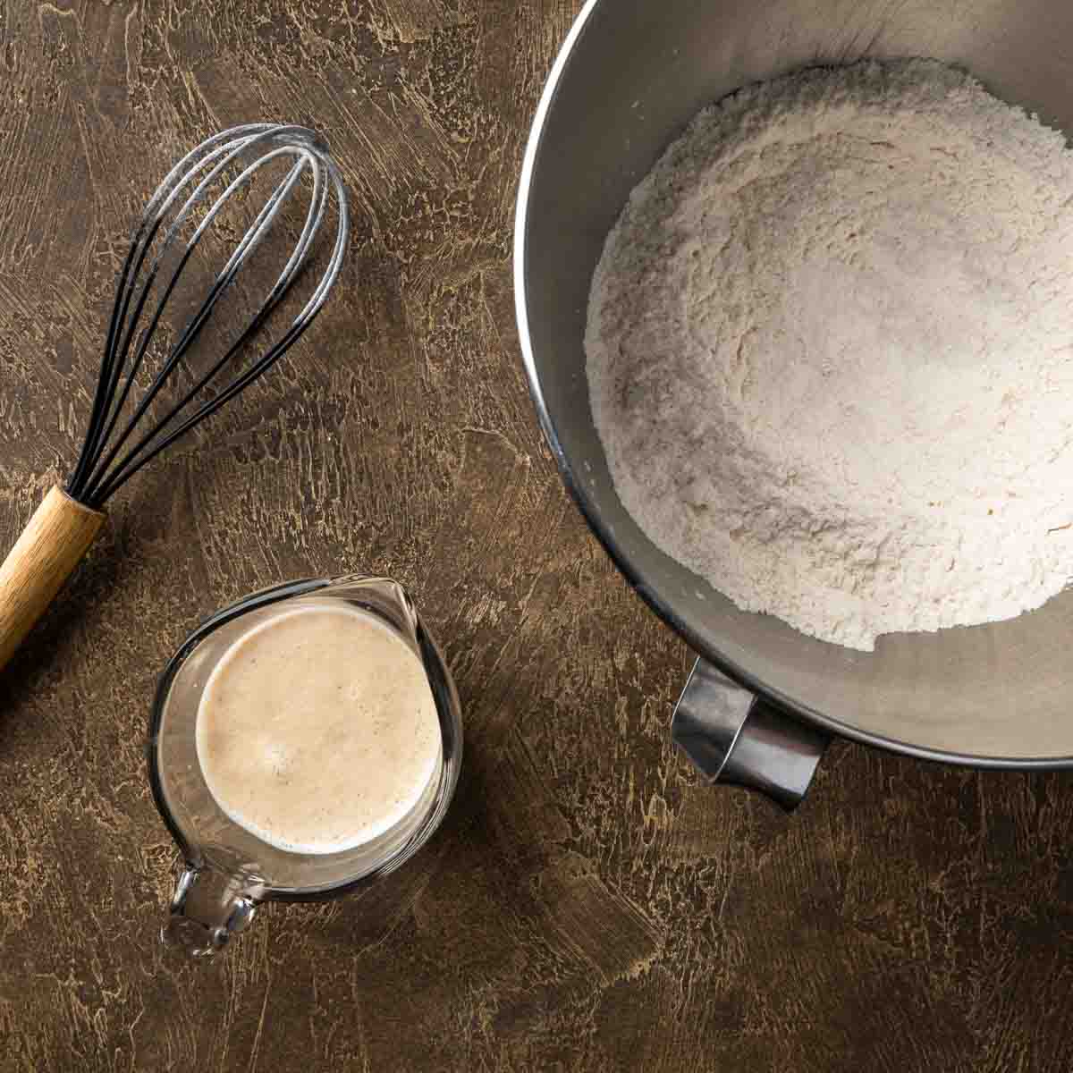 A measuring cup of blooming yeast next to a mixing bowl of whisked flour. 