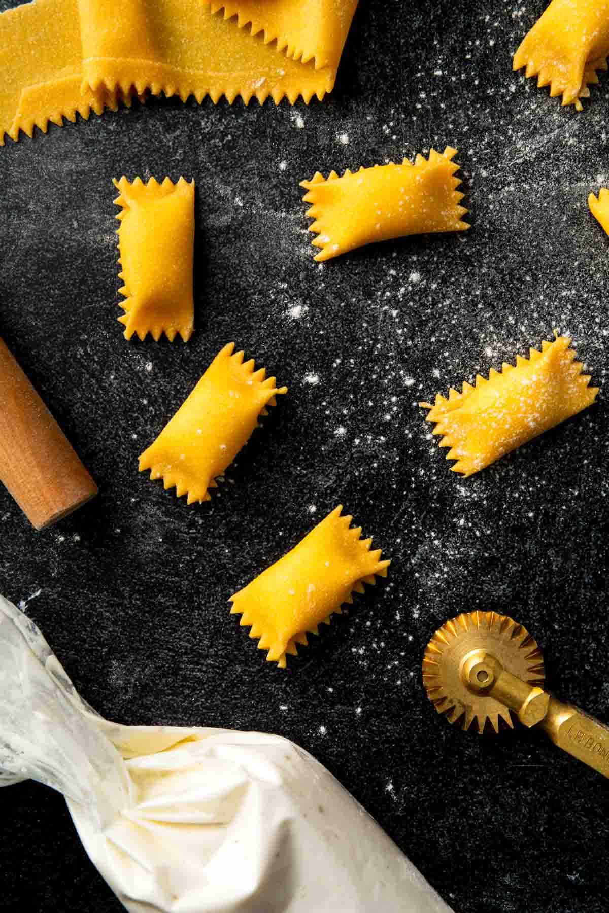 A countertop scene with handmade agnolotti, a ricotta stuffed piping bag and a brass pasta wheel.